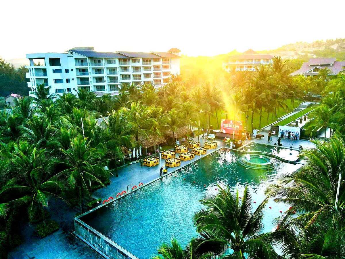 Aerial view of a long blue pool lined with palm trees and buildings, with the morning sun low over the trees