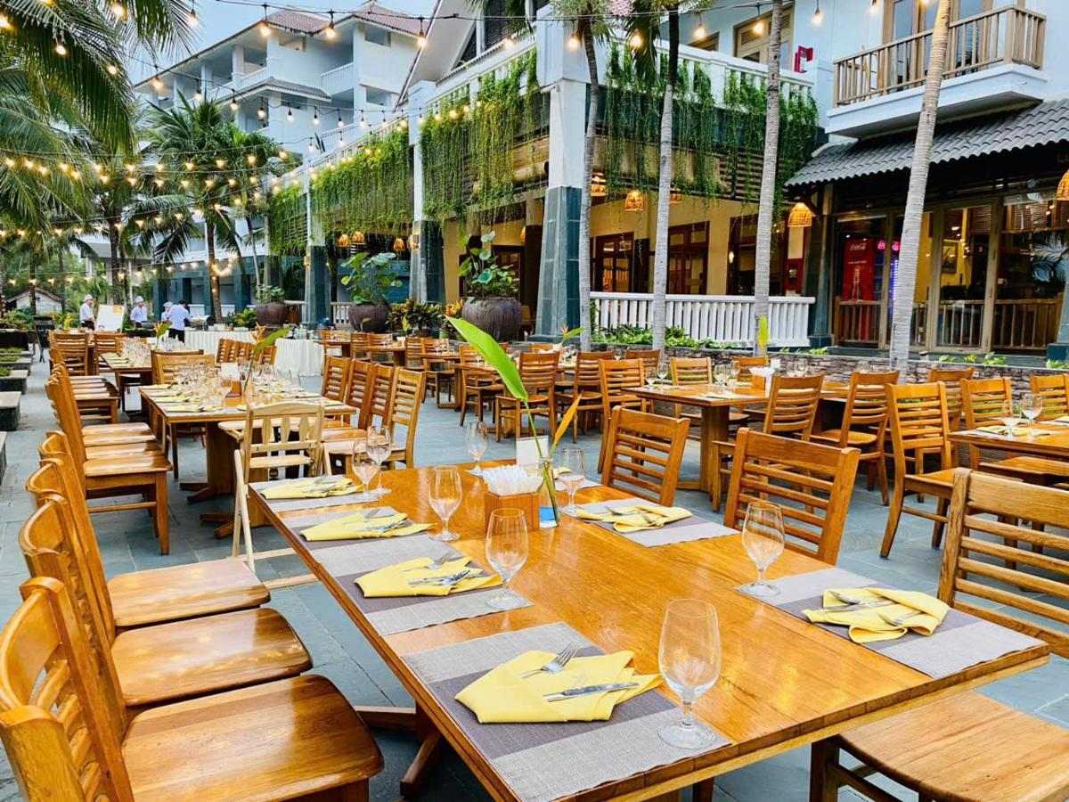 Outdoor restaurant terrace with long wooden tables set with yellow napkins beside a garden