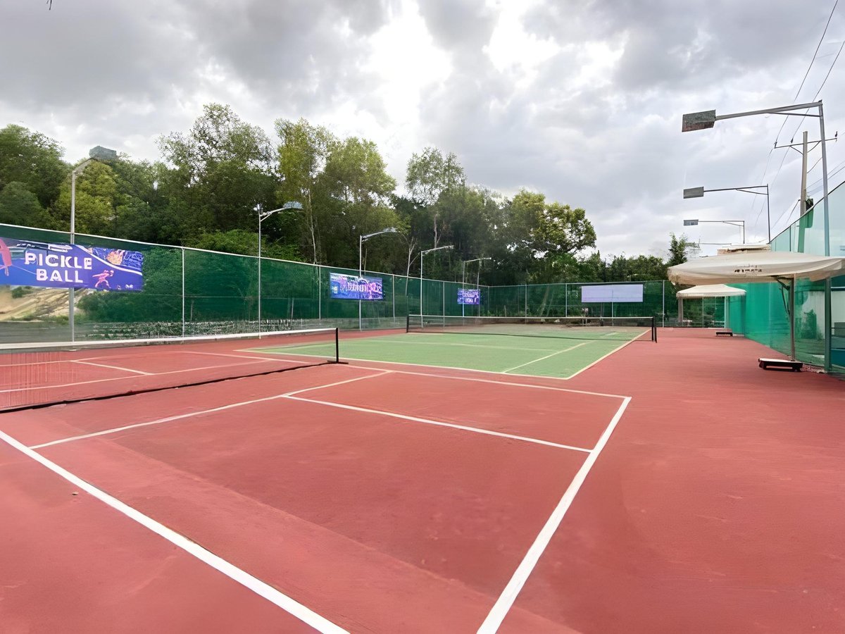 Red and green hard courts with white lines and a net, surrounded by tall green fencing and trees