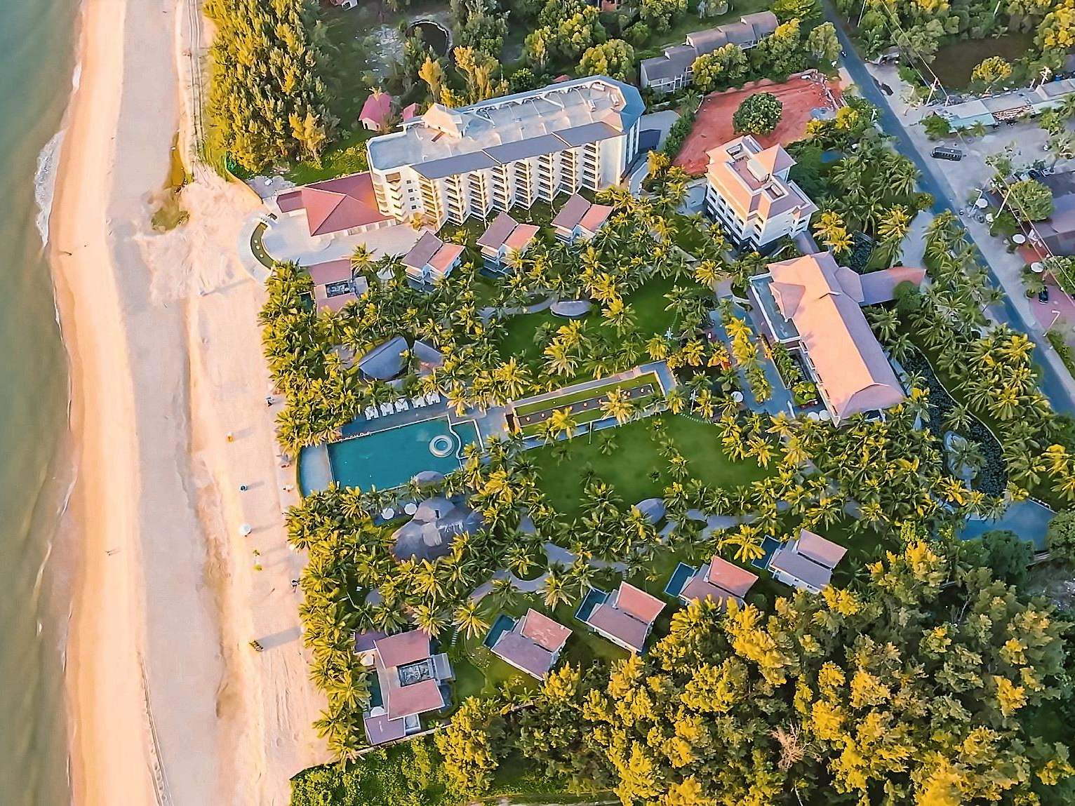 Aerial view of a resort on a coconut grove beside a long beach, with a pool and villas among the palms