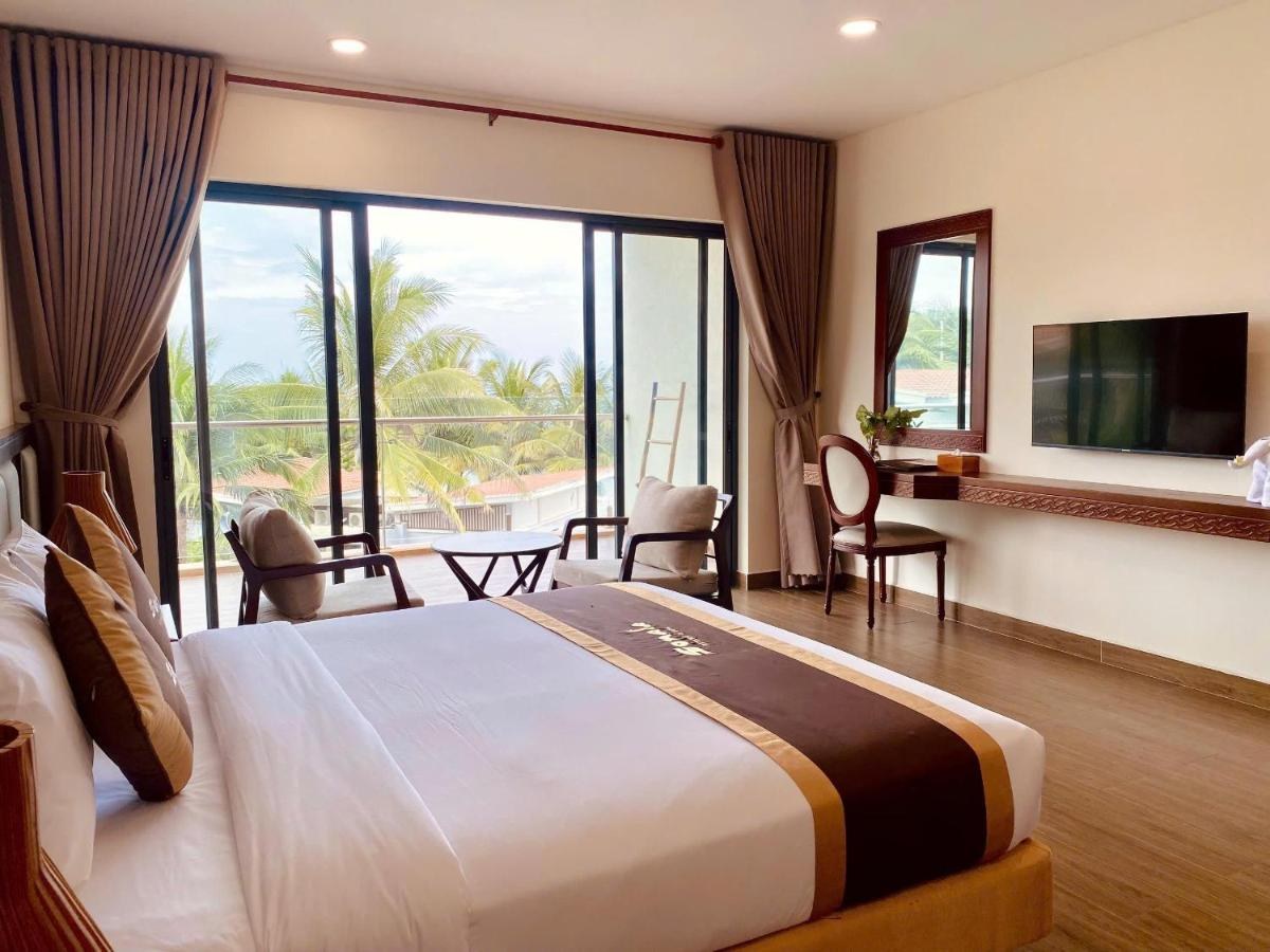 Phòng ngủ có giường đôi, sàn gỗ và cửa kính cao mở ra ban công có ghế, bên ngoài là hàng dừa